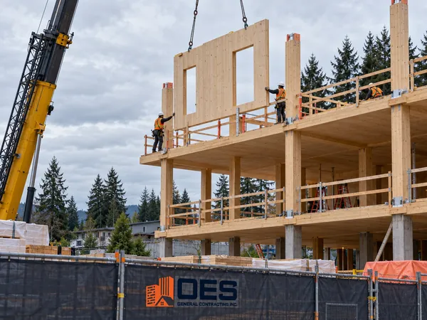 Crane lifting a cross-laminated timber wall panel onto a mass timber frame, with OES site fencing in front