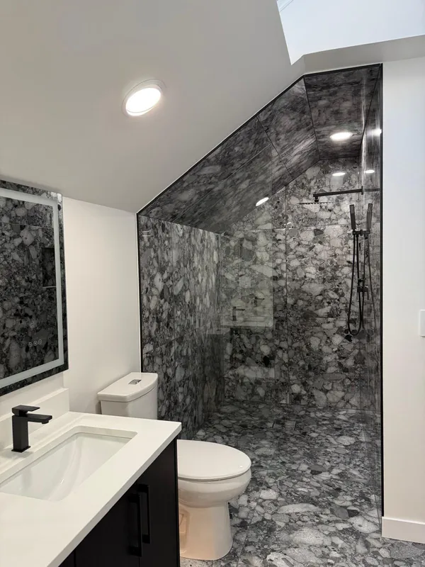 Walk-in shower in large stone-look tile under a sloped ceiling, with a glass panel and black fixtures